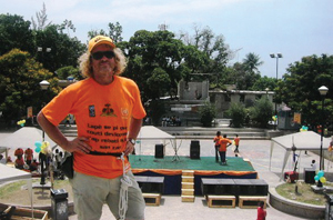Man standing with an orange top and sun glasses on with a stage being set up behind him